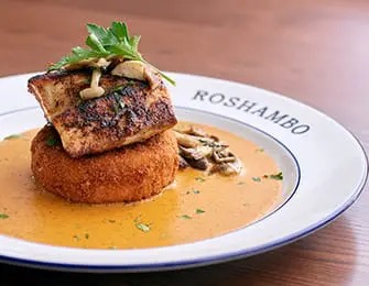 Seared fish fillet on a round fritter with creamy sauce and mushrooms, on a plate with ROSHAMBO