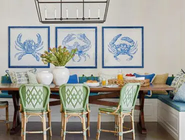 Dining room with banquette seating, wooden table, and green wicker chairs. Blue crab art on wall.