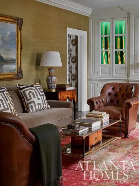 Living room with gold grasscloth wallpaper, brown sofa, leather armchair, and display cabinet with