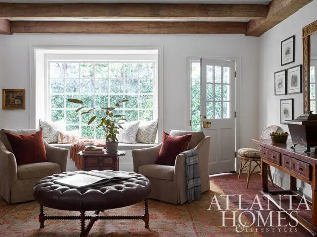 Living room with bay window, tufted leather ottoman, and two armchairs.
