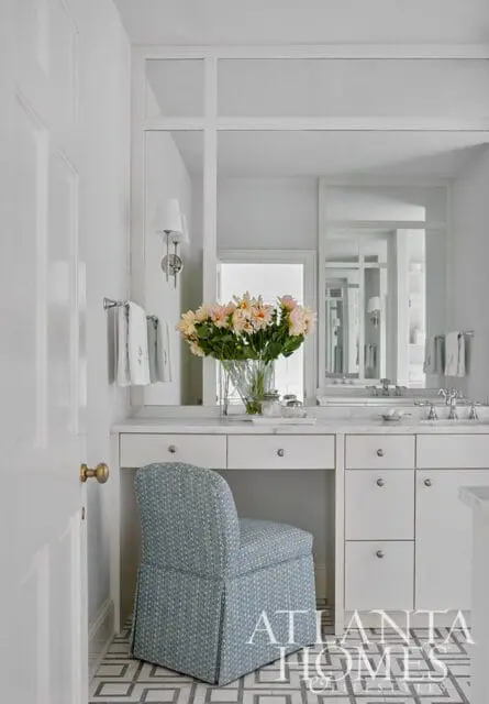White vanity with marble countertop, blue patterned chair, and floral arrangement.
