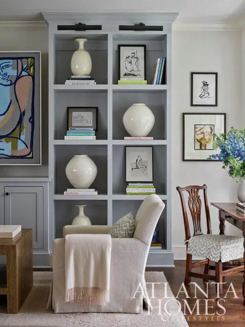 Gray built-in bookshelf with white vases, framed art, and books. Armchair with throw blanket.