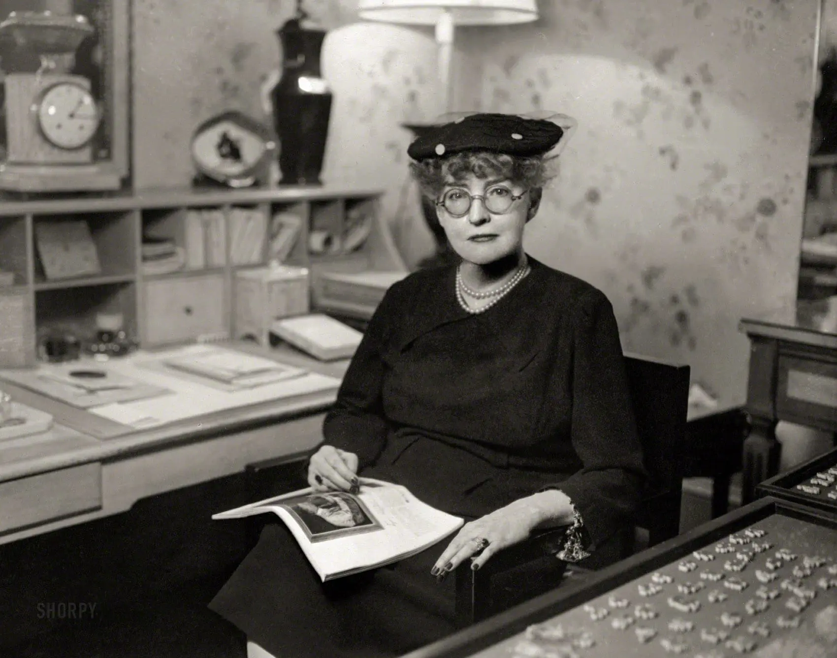 Woman in hat and pearls sitting at desk with magazine.