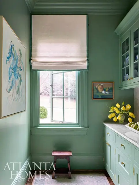 Green kitchen with marble countertops, glass-front cabinets, and a window with a roman shade.