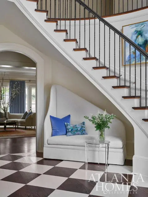 White upholstered bench with blue pillows under a staircase with black spindles.
