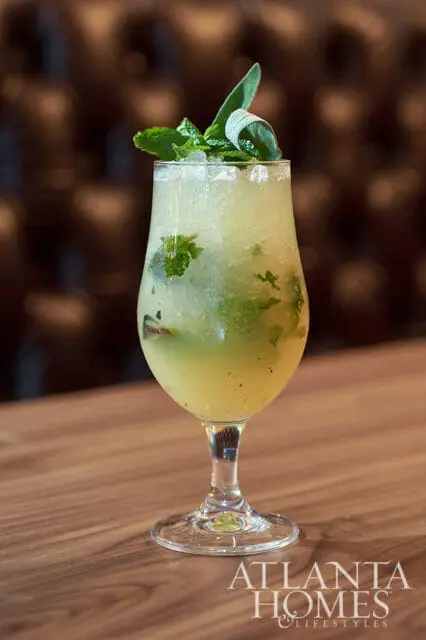 Yellow cocktail with mint and sage garnish in a stemmed glass on a wooden table.