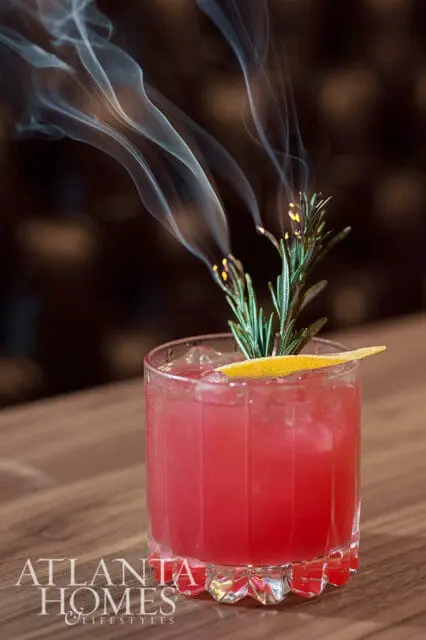 Pink cocktail with ice, orange peel, and smoking rosemary sprig