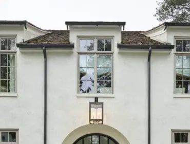 White stucco house with arched black metal door, large windows, and manicured hedges.