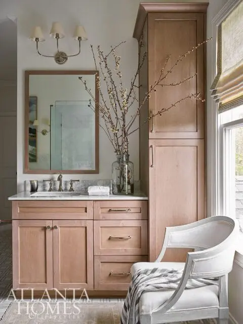 Wood vanity with marble countertop, mirror, and tall cabinet. White chair with striped blanket.