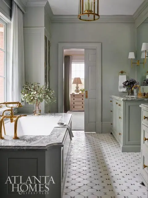 Sage green bathroom with marble tub surround, brass fixtures, and patterned tile floor.