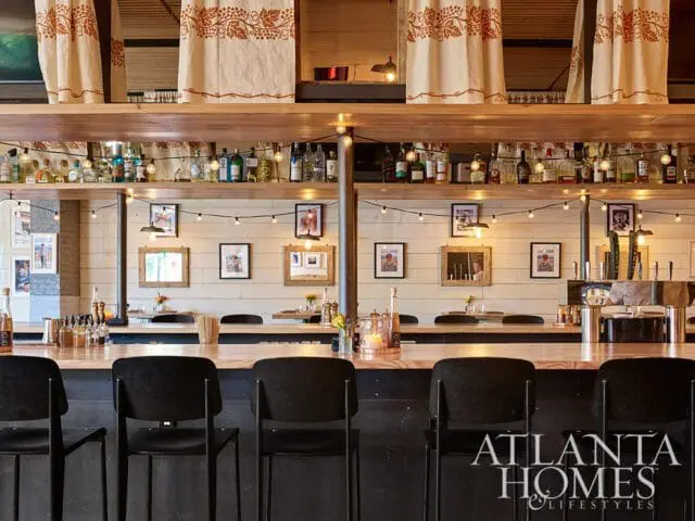 Bar with black stools, wood counter, bottles on shelves, and string lights. Atlanta Homes &