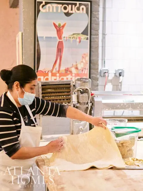 Woman wearing mask making pasta with a machine and vintage poster in background