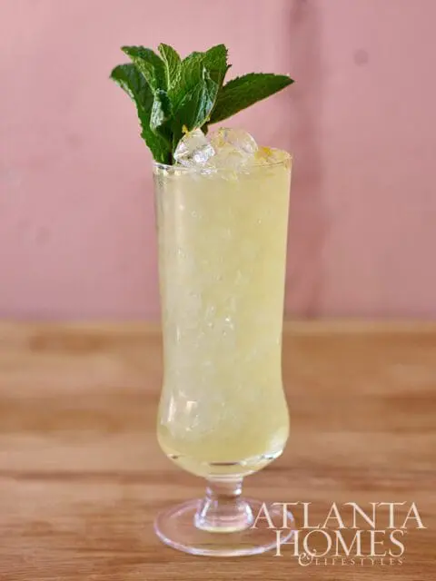 Yellow cocktail with ice and mint garnish in a tall glass with Atlanta Homes & Lifestyles logo