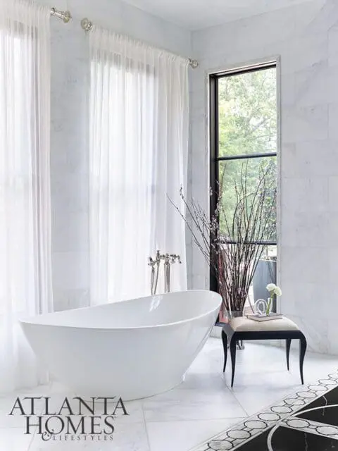White freestanding bathtub in marble bathroom with sheer curtains and tall window