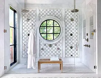 Luxury bathroom with dual showerheads, circular window, marble tile, and wooden bench.