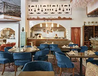 Restaurant dining room with blue velvet chairs, tables set with place settings, and an open kitchen