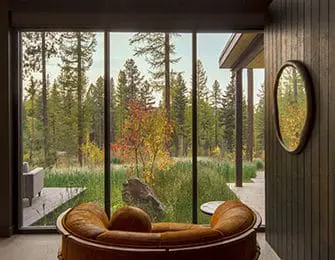 Brown leather sofa facing large window with view of pine forest and colorful autumn foliage.