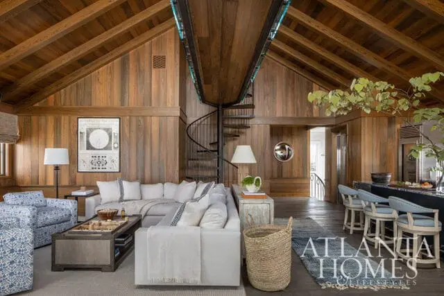 Living room with vaulted wood-paneled ceiling, spiral staircase, sectional sofa, patterned armchairs, and a kitchen island with bar stools.
