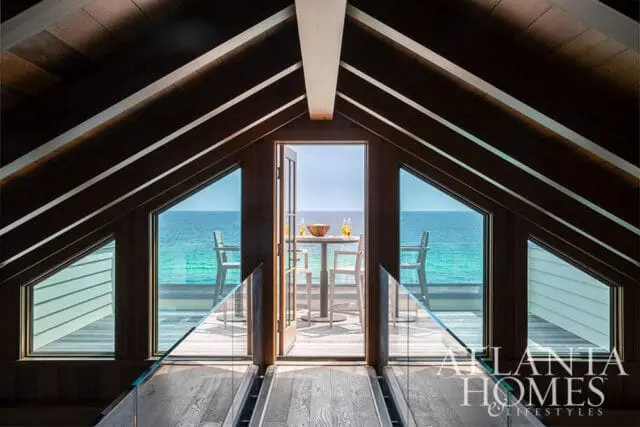 Wooden walkway with glass railings leading to open doors with ocean view, table and chairs on deck