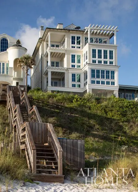 Cream-colored multi-story beach house with balconies and a pergola, accessed by wooden stairs over