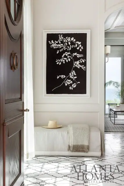 Foyer with dark wood door, white bench with hat and blanket, and framed botanical art.