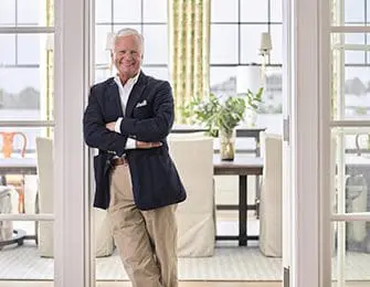 Man in navy blazer and khaki pants smiles with arms crossed in a bright room.
