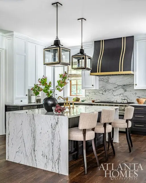 Kitchen island with marble waterfall edge, pendant lights, and black range hood with gold accents.