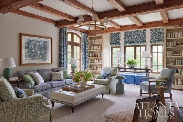 Living room with exposed wood beams, gray sofa, patterned armchairs, and built-in bookshelves.