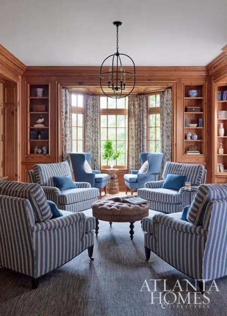 Living room with striped armchairs, blue wingback chairs, tufted ottoman, and wood paneled walls.