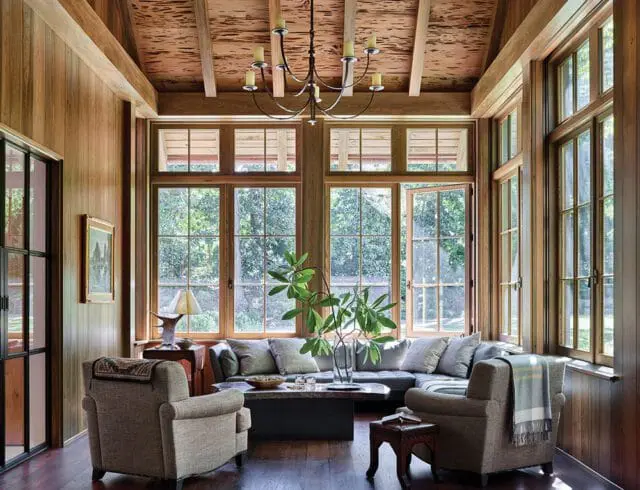 Wood-paneled sunroom with large windows, sectional sofa, armchairs, and a chandelier.