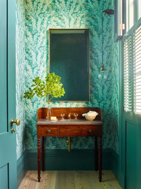 Powder room with teal leaf-patterned wallpaper, antique wood vanity, and brass fixtures.