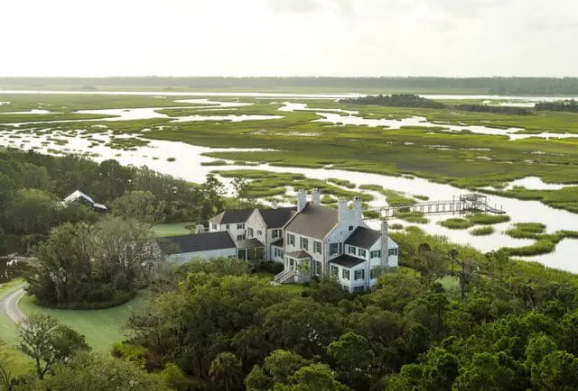 Large white house with green shutters surrounded by trees, overlooking a marsh with a pier.