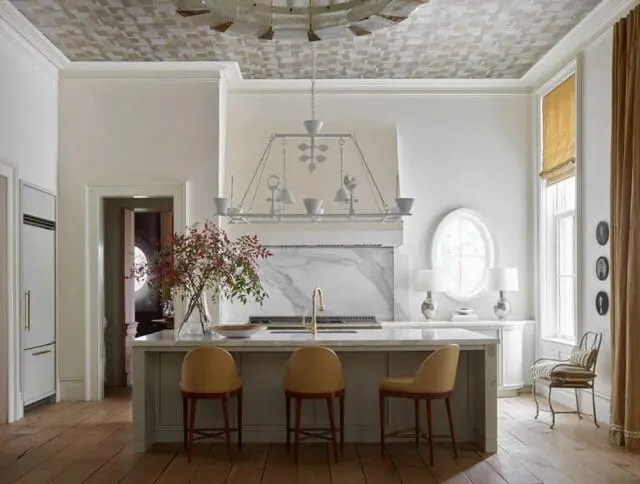Kitchen island with marble countertop and three tan stools, facing a marble backsplash and range