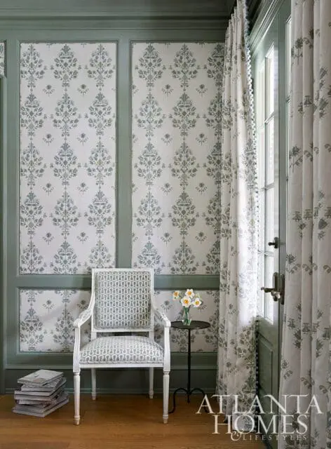 Green paneled walls with damask wallpaper, armchair with geometric pattern, and floral curtains.