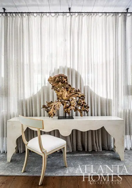Cream console table with ornate gold sculpture and upholstered chair in front of sheer curtains.