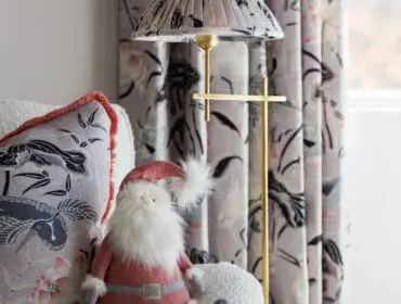 Plush Santa toy on a white boucle sofa next to a patterned pillow and floral curtains.