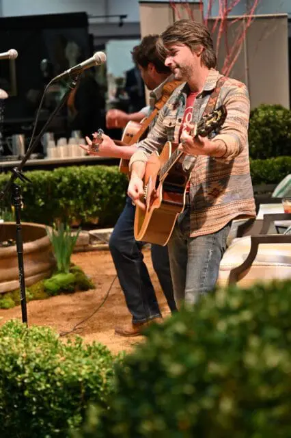 Man in patterned shirt playing acoustic guitar, another man behind him also playing guitar