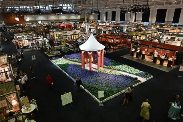 Indoor exhibition hall with numerous booths, a central garden display with purple and white