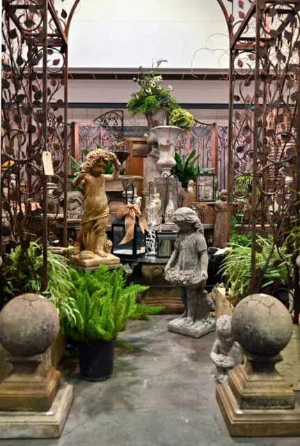 Garden statues and urns displayed under a metal arbor with leafy scrollwork.