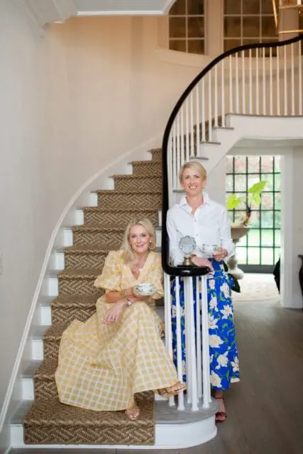 Caroline Cook and Susan Weathersby on a curved staircase holding teacups.