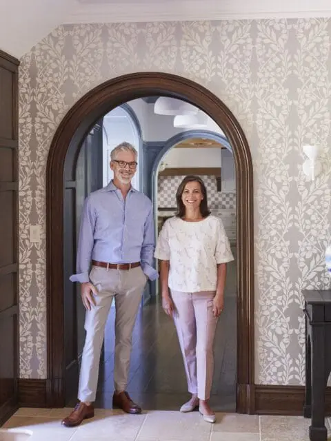 Man in blue shirt and tan pants, woman in white top and pink pants, standing in arched doorway with