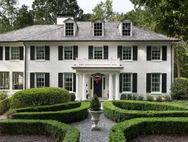 White house with black shutters and dormer windows, surrounded by manicured hedges.