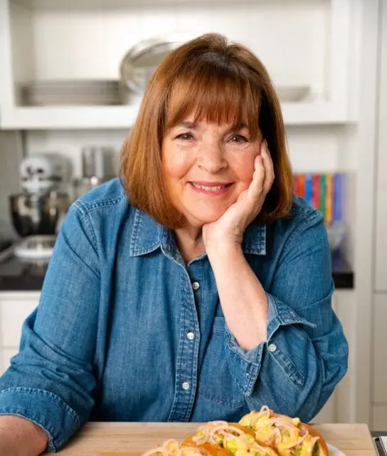 Ina Garten in a denim shirt rests her chin on her hand in a kitchen.