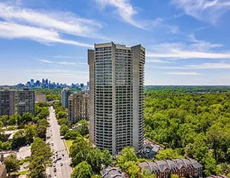 Tall concrete apartment building with many balconies, surrounded by trees and city skyline