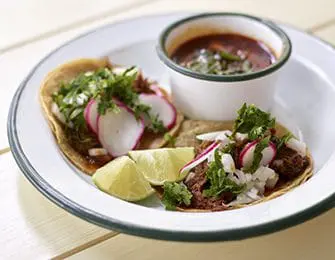 Two tacos with meat, radish, onion, and cilantro, served with a side of broth and lime wedges.