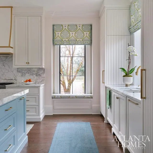 Blue kitchen island with marble countertops and white cabinets. Window has patterned roman shades.