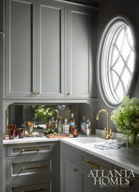 Gray bar area with marble countertop, brass faucet, mirrored backsplash, and oval window.