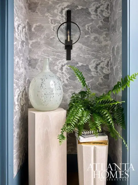 Light wood pedestal with speckled vase, fern on books, and sconce on cloud wallpaper.
