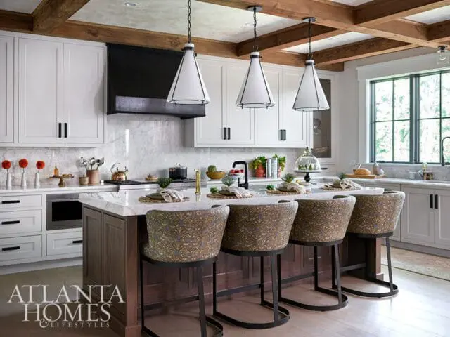 White kitchen with marble island, wood beams, and upholstered bar stools.
