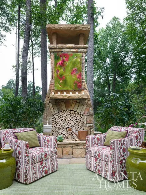 Two armchairs upholstered in floral fabric flank a stone fireplace with stacked firewood and